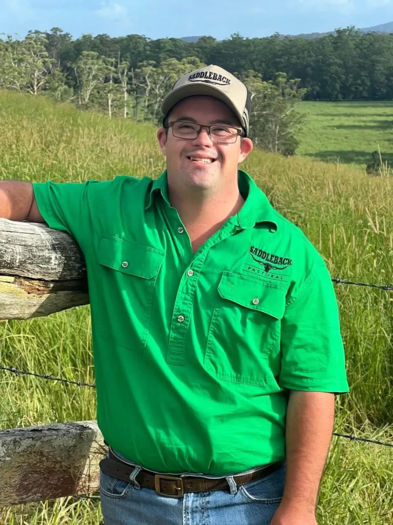 Man in a green Saddleback shirt and cap leaning on a wooden fence in a grassy field with trees in the background.