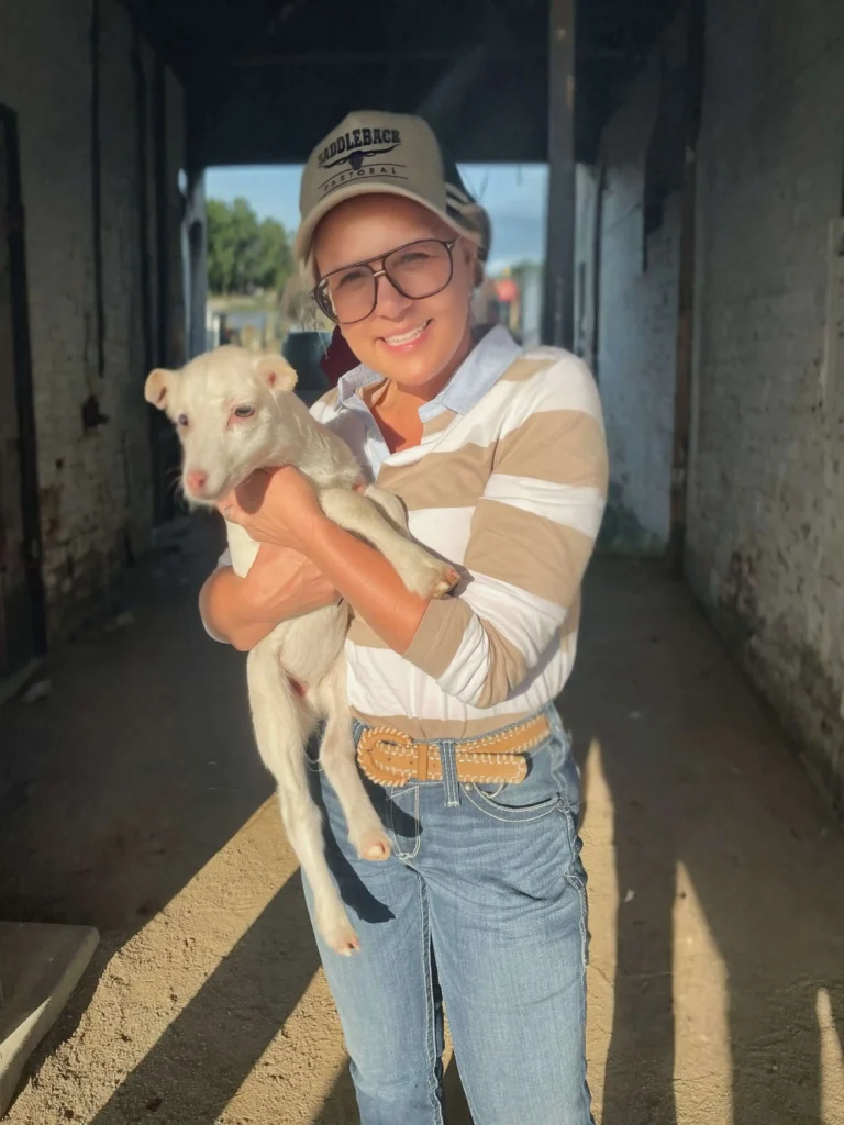 Person holding a small white dog while smiling, standing in a shaded area with sunlight highlighting them. They are wearing glasses, a striped shirt, jeans, and a baseball cap.