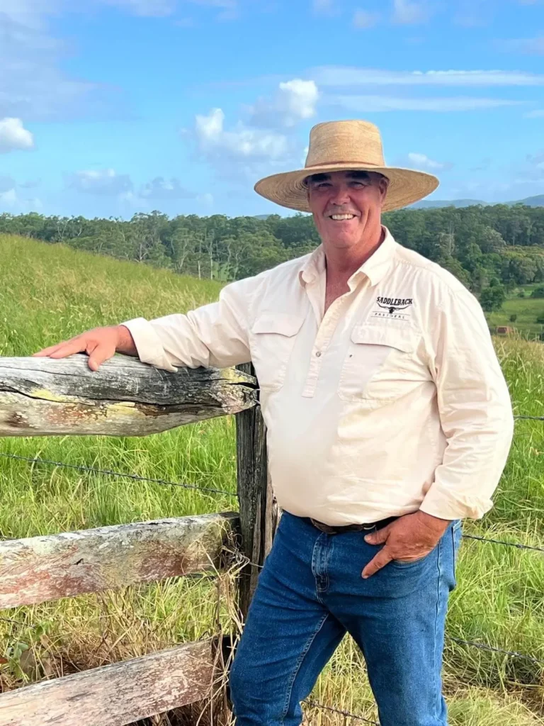 A man in a straw hat and light shirt stands by a wooden fence in a grassy field, with trees and a blue sky in the background.