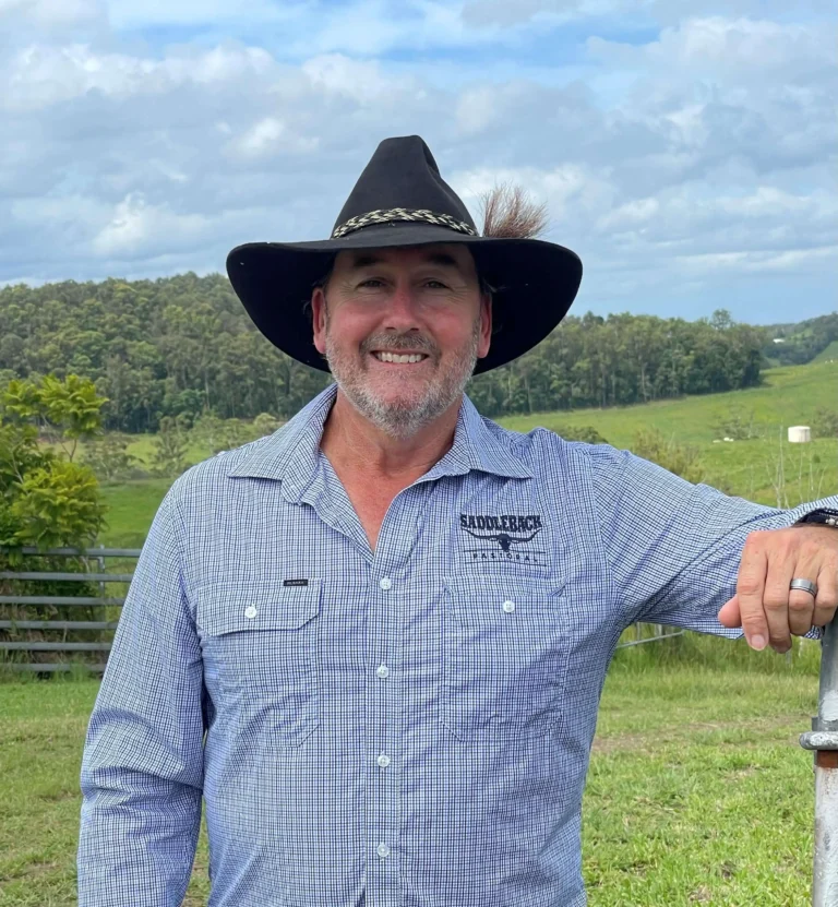 Man wearing a cowboy hat and checkered shirt stands by a gate, smiling, with green hills in the background.