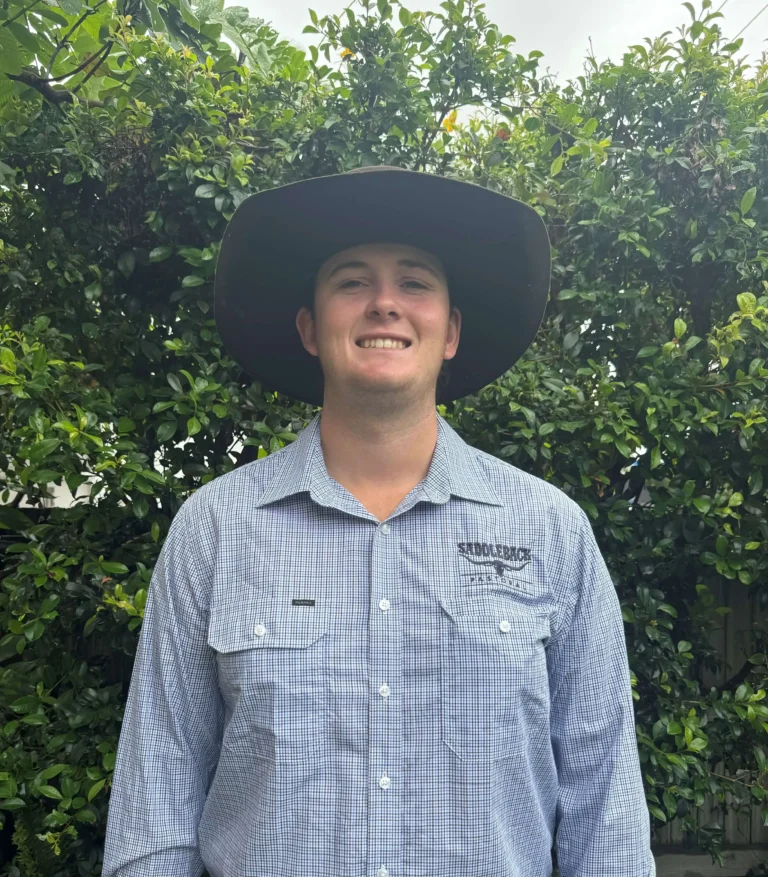 Person wearing a wide-brimmed hat and a checkered shirt stands smiling in front of green foliage.